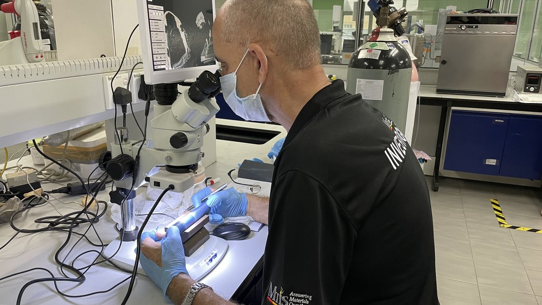 An MTIS engineer re-examining a specimen on a stereo microscope, the live image on the screen beside him